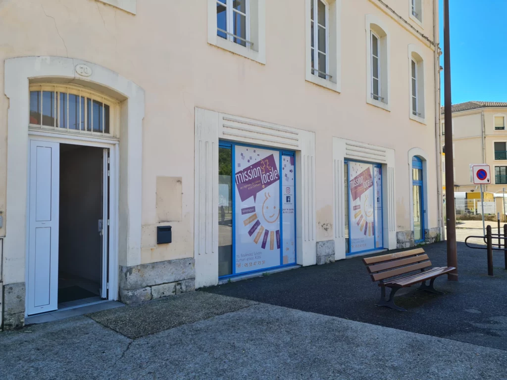 Photo de la façade du batiment de la Mission Locale d'Agen. dans un vieux batiment de couleur beige/rosé, avec des entouragages de pierre nous avons une porte blanche, 2 vitrine et une porte bleue.