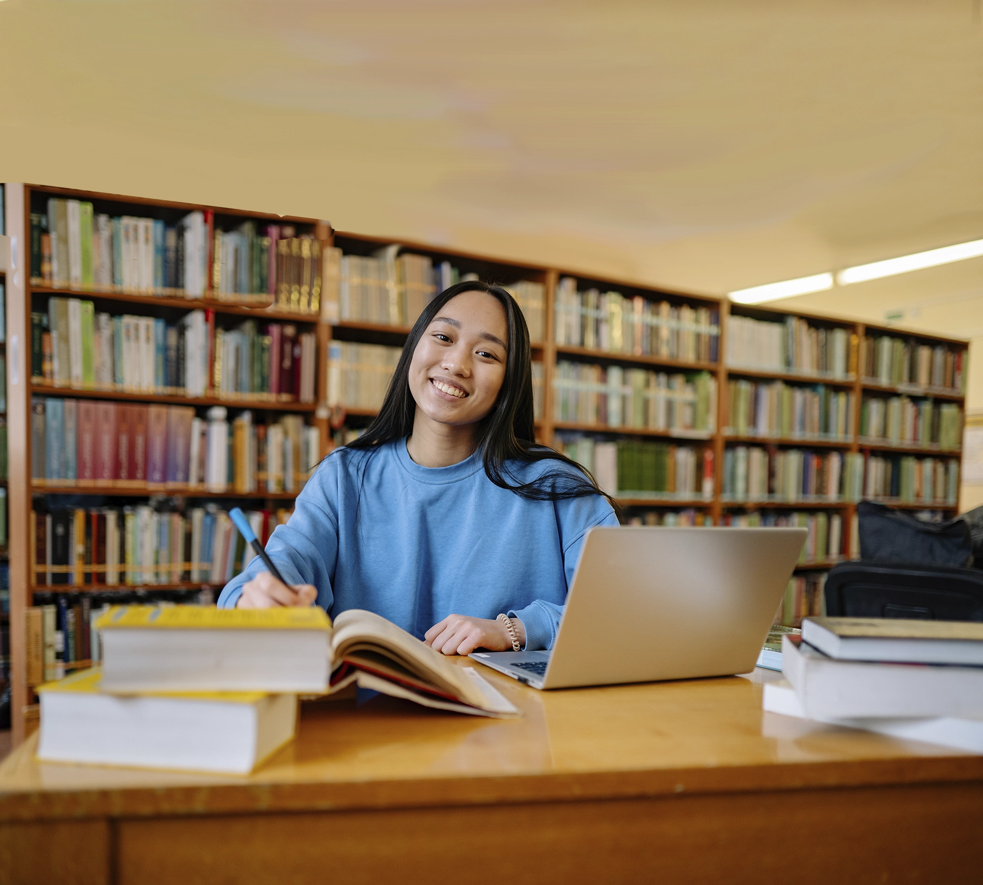 Dans une bibliothèque, posée à un bureau, une jeune femme aux cheveux bruns étudie avec pleins de livres et un ordinateur portable.