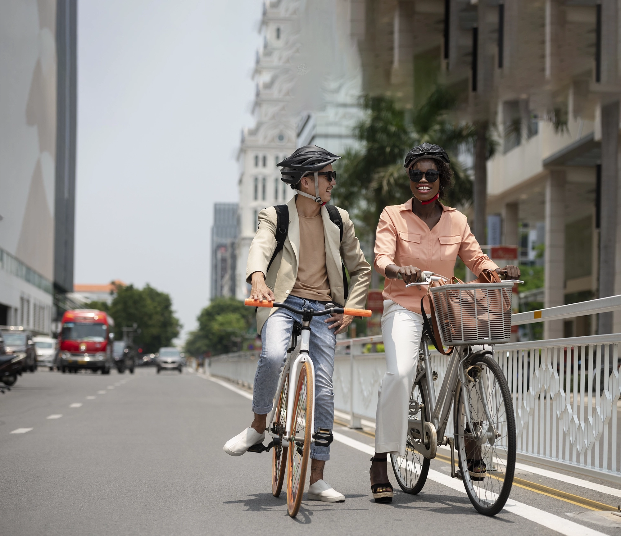 Dans une rue un homme caucasien et une femme africaine discutent chacun sur un vélo. Ils ont tous les deux des casques de sécurité et des lunettes de soleil. L'homme a un vélo blanc avec des roue oranges et la femme un vélo gris avec un panier dans lequel est posé son sac à main.